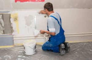 A man in blue overalls performing water damage restoration in San Diego, CA, using a roller to repair and restore a water-damaged wall.