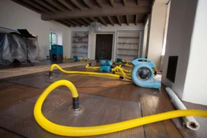 pipe water cleanup Industrial water damage restoration equipment, including blue air movers and yellow suction hoses, being used to dry a dark hardwood floor in a room with protective plastic coverings.